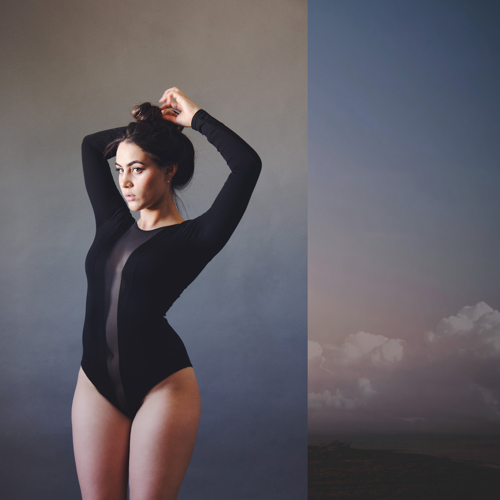 Valeria Cisneros poses elegantly in a black bodysuit against a muted gray backdrop with her hands gracefully holding her hair juxtaposed against a serene landscape of a cloudy sky and distant hills.